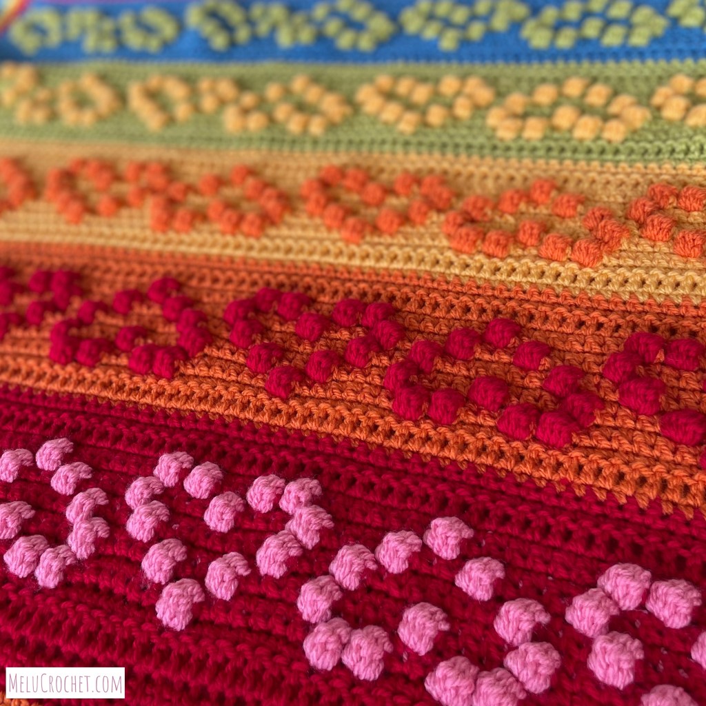Rainbow coloured blanket with raised bumps that are formed using the crochet bobble stitch in the shape of hearts fading out of focus in the distance