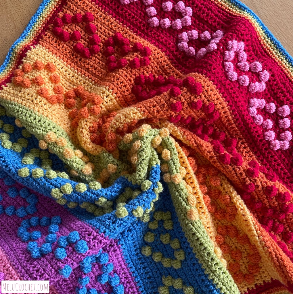 Rainbow coloured blanket with raised bumps that are formed using the crochet bobble stitch in the shape of hearts. The blanket has been swirled.