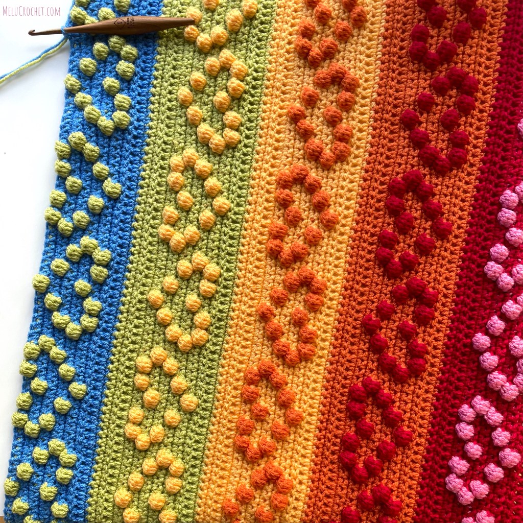 Progress photo of Rainbow coloured blanket with raised bumps that are formed using the crochet bobble stitch in the shape of hearts, with furls crochet hook