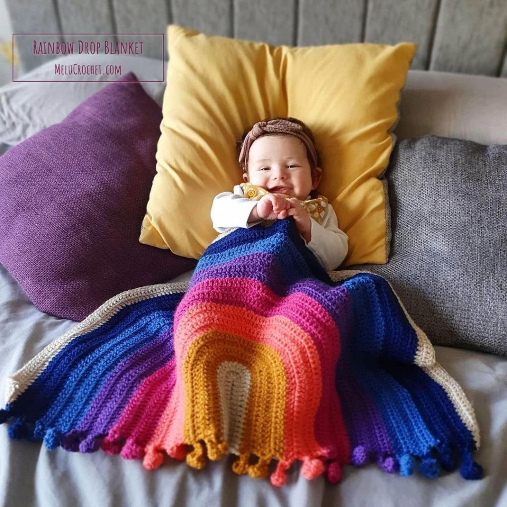 Cute smiling Baby girl snuggled with her crocheted rainbow drop blanket