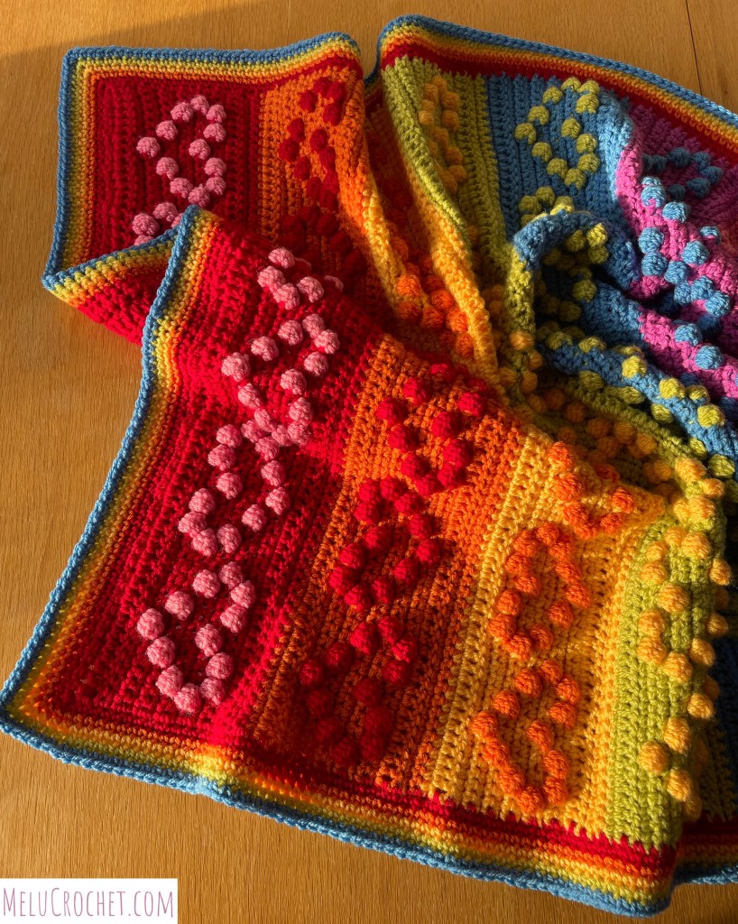 Rainbow coloured blanket with raised bumps that are formed using the crochet bobble stitch in the shape of hearts, crumpled up and in sunshine