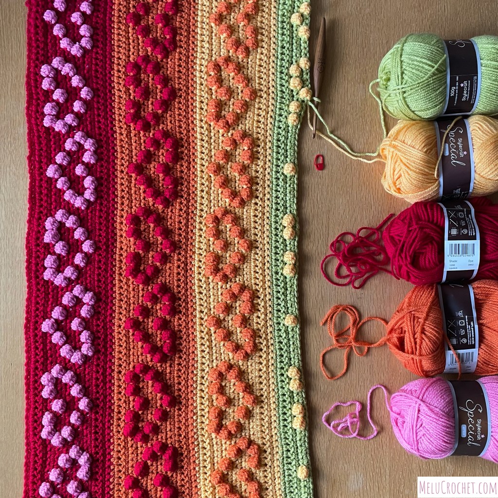 Construction of a Rainbow coloured blanket with raised bumps that are formed using the crochet bobble stitch in the shape of hearts, each colour has a ball of stylecraft wool that is being used, along with a furls crochet hook